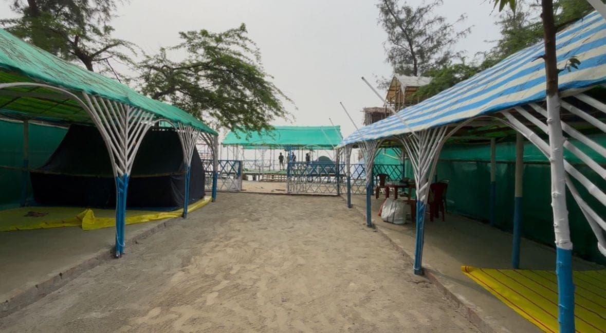 A sand path between shaded outdoor structures at the camp, with water in the distance